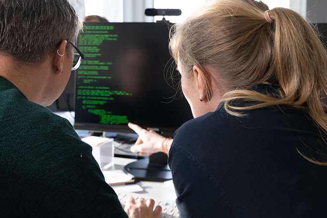 Teaserbild zeigt zwei Mitarbeitende vor einem Monitor mit Code.